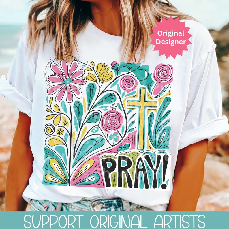 May include: White t-shirt with a colorful floral design, a yellow cross, and the word "PRAY!" in black. The design includes pink, blue, yellow, and green elements. The shirt has rolled-up sleeves.