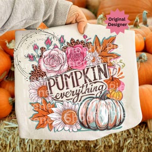 May include: A cream-colored tote bag featuring a floral and pumpkin design. The design includes pink roses, orange leaves, sunflowers, and the words "PUMPKIN everything". The bag is held in front of a background of pumpkins.