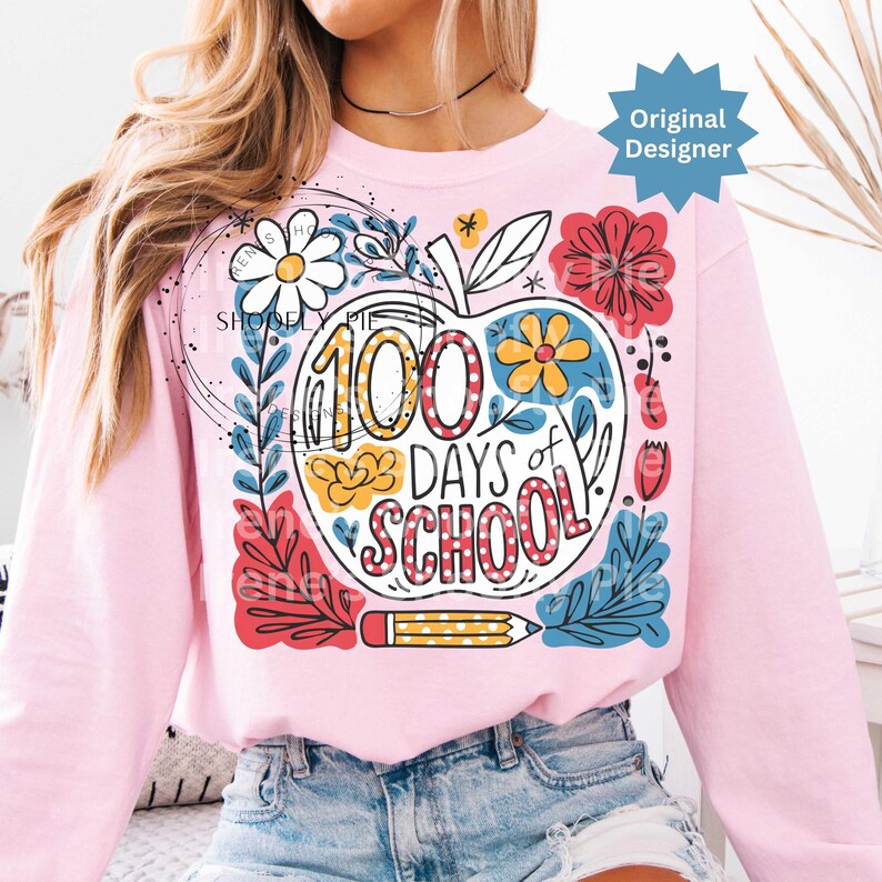 May include: A pale pink sweatshirt with a vibrant apple graphic. The apple design includes the text "100 Days of School", flowers, and a pencil. The sweatshirt is a long-sleeved crew neck.
