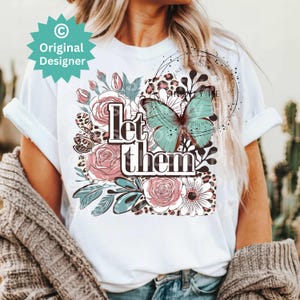 May include: White t-shirt with a floral design, a butterfly, and the text "Let them" printed on the front.