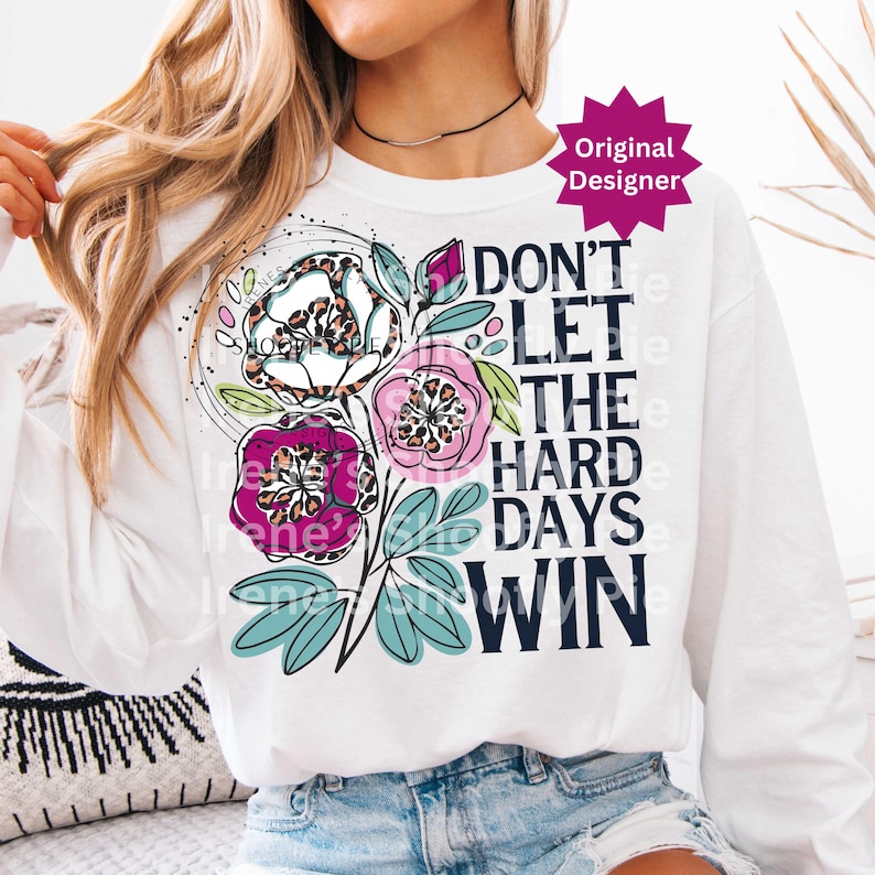 Op de afbeelding: Wit sweatshirt met lange mouwen met een bloemenontwerp en de tekst "DON'T LET THE HARD DAYS WIN". Het bloemenontwerp bevat roze, paarse en witte bloemen met luipaardprint accenten en blauwgroene bladeren.
