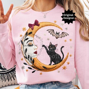 Op de afbeelding: Een lichtroze sweatshirt met een geborduurd ontwerp van een maan, een zwarte kat en een vleermuis. De maan is goudkleurig met een bordeauxrode strik. Het ontwerp bevat sterren en de tekst "SHOOFY PIE DESIGN".