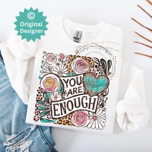 May include: White t-shirt with a floral design and the text "You are Enough" in black and teal lettering. The design includes a heart with a teal and white pattern.