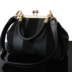 Peut inclure: Sac à main noir avec un design structuré. Le sac est doté d'une poignée supérieure arrondie et d'une bandoulière amovible. Des éléments dorés ornent le sac, notamment la fermeture à fermoir et les attaches de la bandoulière.