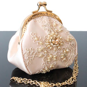 Peut inclure: Une pochette en satin rose pâle dotée d'un fermoir à cliquet en métal doré et d'une bandoulière en chaîne dorée amovible. L'avant est orné de broderies florales en dentelle blanche, de petites perles crème et d'un cristal couleur champagne.
