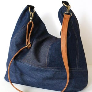 Peut inclure: Sac à bandoulière en jean bleu foncé avec une bandoulière en cuir marron et des accessoires métalliques dorés. Le sac présente un design cousu. La bandoulière est réglable et peut être portée à l'épaule ou en travers du corps.