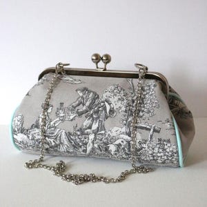 Peut inclure: Une pochette en tissu toile de Jouy gris et blanc représentant une scène pastorale avec des personnages, des arbres et une cage à oiseaux. Le sac possède un cadre en métal argenté avec un fermoir à deux boules, une chaîne amovible et un passepoil bleu clair.