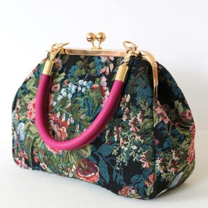 Peut inclure: Sac à main de style vintage avec un motif de tapisserie florale. Le sac présente un fond noir avec un motif coloré de fleurs dans les tons de rose, de bleu et de vert. Il a un fermoir doré et des poignées roses.