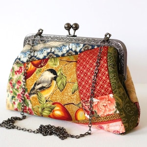 Peut inclure: Une pochette en tissu patchwork ornée d'un motif d'oiseau et de fruits dans des tons jaune, rouge et vert. Le sac possède une armature en métal foncé avec un fermoir à deux boules et une bandoulière en chaîne métallique amovible.