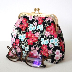 Peut inclure: Un porte-monnaie en tissu noir orné d'un motif floral vibrant de fleurs de cerisier rouges, roses et bleu clair. Le porte-monnaie est doté d'une fermeture à fermoir métallique doré. Une paire de lunettes à monture brune repose devant.