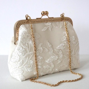 May include: Cream-coloured clutch bag with a gold-tone clasp and chain strap. The fabric is decorated with white floral embroidery. The clasp has a decorative design. A classic accessory for special occasions.