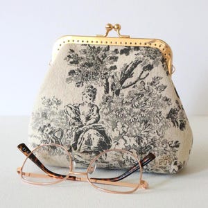 Peut inclure: Un porte-monnaie en tissu beige avec un imprimé toile de jouy noir représentant une silhouette assise et des arbres, doté d'un fermoir à cliquet en métal doré. Devant, une paire de lunettes à monture fine dorée et branches écaille.