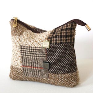Peut inclure: Une pochette en tissu patchwork avec une fermeture éclair marron et une tirette dorée. La pochette présente une variété de motifs tweed dans les tons marron, beige et noir, avec deux boutons carrés décoratifs.