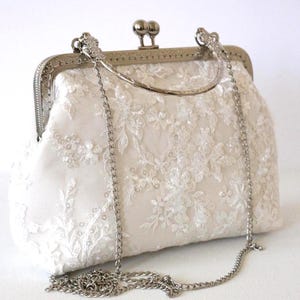Peut inclure: Une pochette couleur crème ornée d'une superposition de dentelle florale blanche avec de subtiles paillettes. Le sac présente un cadre en métal argenté, une fermeture à fermoir, une courte poignée incurvée et une bandoulière en chaîne argentée.