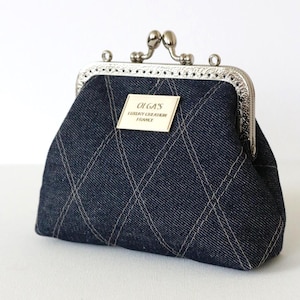 Peut inclure: Un porte-monnaie en jean bleu foncé avec un fermoir et un cadre argentés. Le sac à main présente un motif matelassé avec des coutures claires. Une petite étiquette rectangulaire indique "OLGA'S LUXURY CREATION FRANCE".