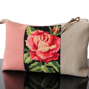 Peut inclure: Une trousse de maquillage rectangulaire avec un motif floral. Le sac présente une grande rose brodée dans les tons de rouge, rose et vert sur fond noir. Le sac est en tissu rose et beige avec une fermeture éclair.