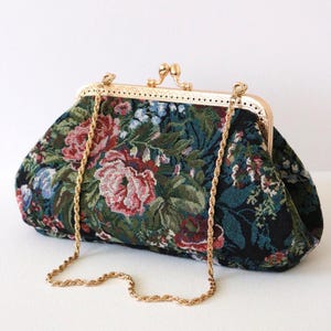 Peut inclure: Une pochette structurée avec une base en tissu sombre ornée d'un motif floral tissé dans des tons de rouge, vert et bleu. Le sac possède un cadre en métal doré avec un fermoir à pression et une fine bandoulière en chaîne métallique torsadée.