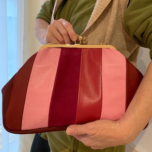 Peut inclure: Un sac à main de type pochette avec un fermoir doré. Il présente des rayures verticales de différentes nuances de rouge et de rose. La bande centrale est rose texturée, flanquée de panneaux rouges et bordeaux unis.
