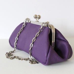 May include: A purple clutch purse with a silver chain strap. The purse has a kiss-lock closure and a textured leather exterior. The chain is detachable, allowing the purse to be used as a clutch.