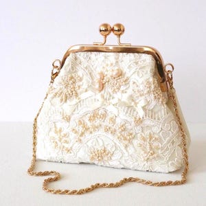 May include: A cream-colored clutch bag featuring intricate white lace overlays, delicate beadwork, and small satin ribbon bows. The bag includes a polished gold-tone metal frame with a dual-ball clasp closure and a matching gold-tone chain shoulder strap.