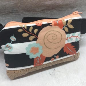 May include: A floral print fanny pack with a black elastic strap. The pack features a black and white striped pattern with peach, gold, and teal floral accents. The bottom is made of cork, and the zipper is peach.