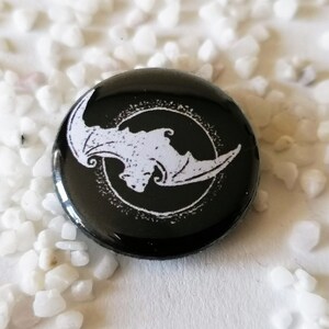 May include: A black button with a white bat design inside a circle. The bat is facing left with its wings spread. The button is glossy and set against a background of small white stones.