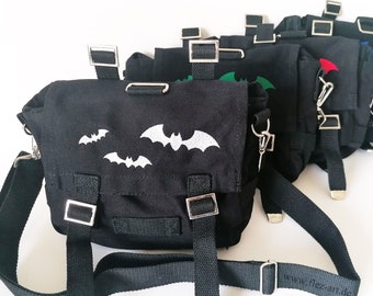 May include: A set of six black canvas bags with adjustable straps. Each bag features a white embroidered bat design and a silver metal buckle. The bags are lined up in a row, with the straps of the bags in the back of the row overlapping the straps of the bags in the front of the row. The bags are all the same size and shape. The text "www.fliezan.de" is visible on the strap of the bag in the front of the row.
