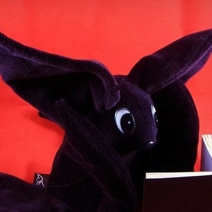 May include: A plush, dark purple bunny toy with large ears and white eyes, reading a small, dark brown book. The bunny is set against a vibrant red background, creating a striking contrast.