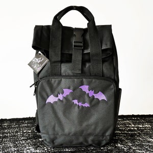 Può includere: Zaino nero con un designo di pipistrello viola. Lo zaino ha una patta con chiusura a fibbia e una tasca con cerniera sul davanti.