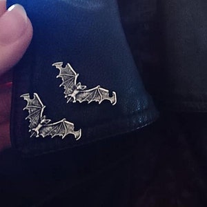May include: Three silver bat pins on a black leather jacket. The bats are in a flying position with their wings spread out.