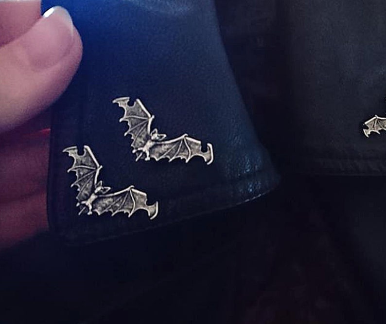 May include: Three silver bat pins on a black leather jacket. The bats are in a flying position with their wings spread out.
