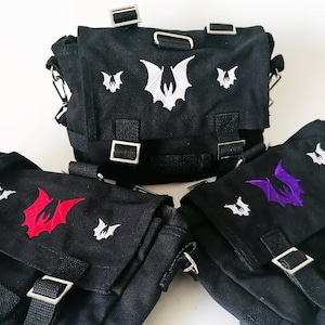 Può includere: Tre borse in tela nera con cinturini regolabili. Ogni borsa presenta un diverso design di pipistrello ricamato: bianco, rosso e viola.
