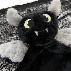 May include: A black plush bat with grey wings and a toothy grin. The bat has large yellow eyes and is sitting on a black spiderweb.