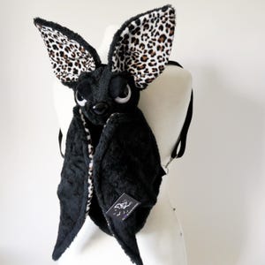 May include: A plush black bat-shaped backpack featuring large, upright ears with a brown and black leopard print interior. The backpack has a soft, fuzzy texture, large white and black eyes, a black nose, and black adjustable shoulder straps.