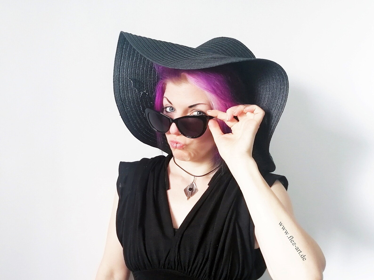 Goth woman in wide-brim sun hat and black dress with purple hair and sunglasses, channeling playful summer energy.