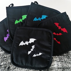 Op de afbeelding: Set van vijf zwarte ovenwanten met geborduurde vleermuizen in verschillende kleuren, waaronder wit, rood, groen, blauw en paars. De ovenwanten zijn gemaakt van een zachte, gewatteerde stof en hebben een lus om op te hangen.