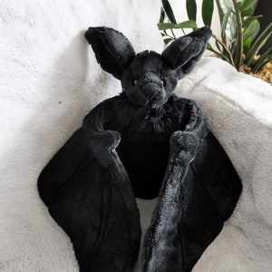Könnte beinhalten: Eine schwarze Plüschfledermaus mit großen Flügeln. Die Fledermaus sitzt auf einer weißen, flauschigen Oberfläche.