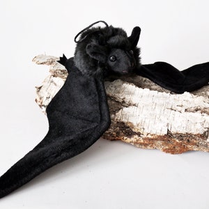 May include: A black plush bat with large wings, sitting on a piece of birch bark.