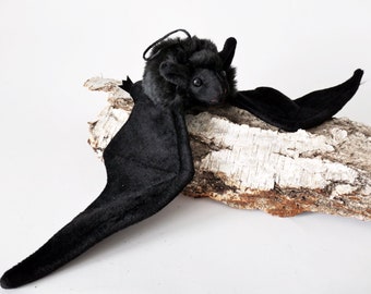 Könnte beinhalten: Eine schwarze Plüschfledermaus mit großen Flügeln, die auf einem Stück Birkenrinde sitzt.