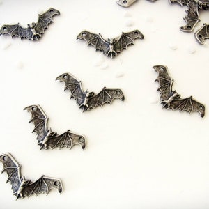May include: A collection of silver-toned metal bat charms with detailed wings and a small bat figure in the center. The charms have two holes for attaching to jewelry or other crafts.