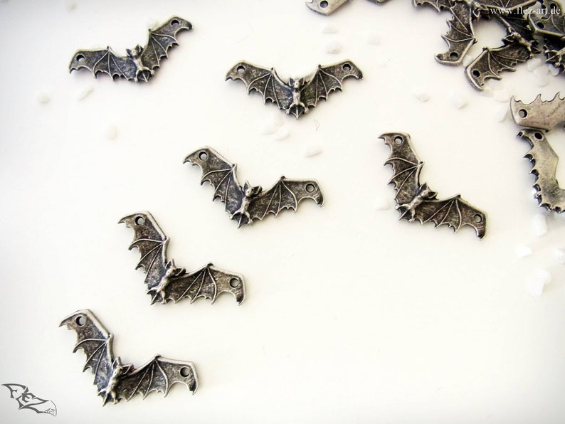 May include: A collection of silver-toned metal bat charms with detailed wings and a small bat figure in the center. The charms have two holes for attaching to jewelry or other crafts.