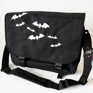 Può includere: Borsa a tracolla nera con silhouette di pipistrelli bianche ricamate sul davanti. La borsa ha una lunga tracolla regolabile e una chiusura a lembo con due fibbie.