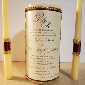 May include: Wedding unity candles with a burgundy ribbon and gold accents. The candle label has the text "R&A" and the names "Rebecca Marie" and "Alfred Joseph Yakaboski" with the date "Saturday, the Fourteenth of August Two Thousand Twenty-One" and the time "At Two Thirty in the Afternoon".