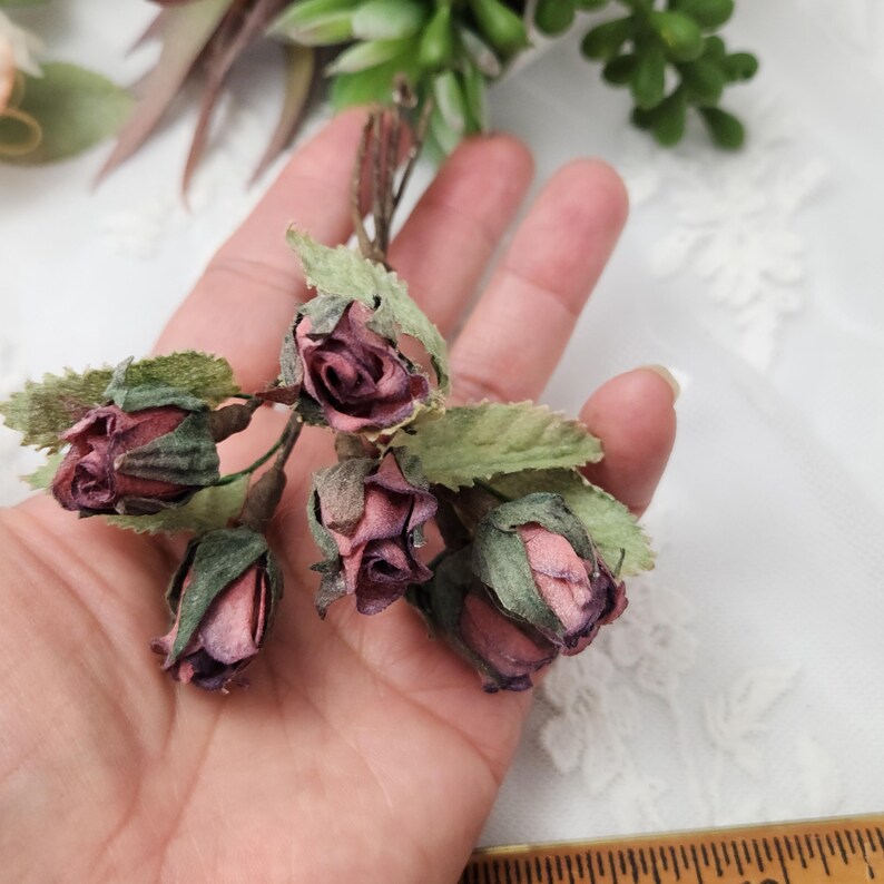 Pu&ograve; includere: Primo piano di un gruppo di piccole rose artificiali in varie tonalit&agrave; di rosa e viola, con foglie verdi. Le rose sono raggruppate e sembrano essere fatte di carta o tessuto. L'estetica generale &egrave; delicata e vintage.