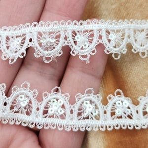 May include: White lace trim with small white sequins. The trim has a scalloped edge and is perfect for adding a touch of elegance to any project.