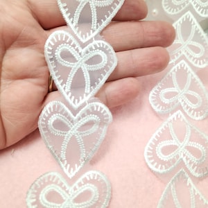 May include: White lace trim with a repeating pattern of hearts with bows. The lace is sheer and delicate.
