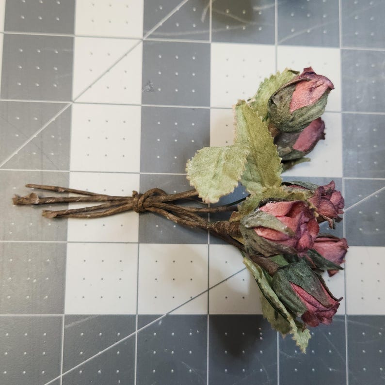 Pu&ograve; includere: Un piccolo mazzo di rose secche con petali rosa e bordeaux e foglie verdi. Gli steli sono legati con spago. Le rose sono disposte su una superficie a griglia grigia e bianca.