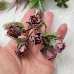 Pu&ograve; includere: Primo piano di un rametto di rose in miniatura fatte a mano. Le rose sono di un colore bordeaux intenso con riflessi rosa e le foglie sono verdi. Le rose sono disposte su uno stelo marrone.