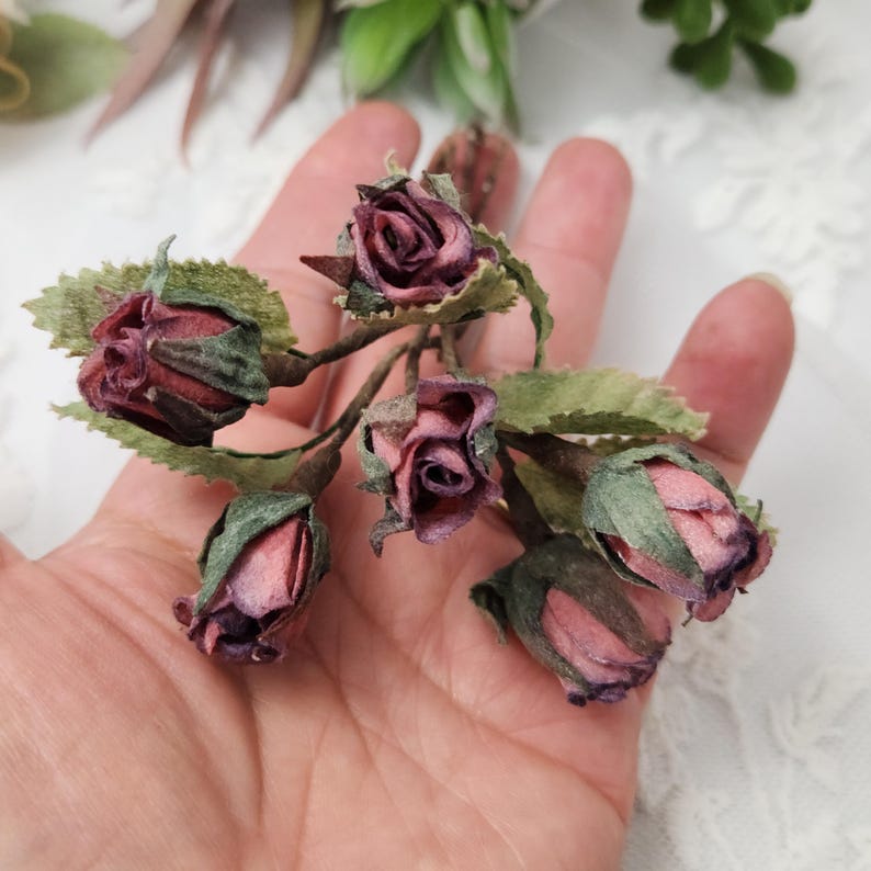 Pu&ograve; includere: Primo piano di un rametto di rose in miniatura fatte a mano. Le rose sono di un colore bordeaux intenso con riflessi rosa e le foglie sono verdi. Le rose sono disposte su uno stelo marrone.