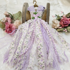 May include: A miniature purple and white floral dress with lace trim and a pearl necklace, hanging on a wooden spool.